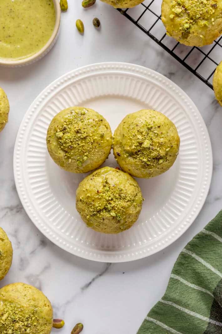 Pistachio muffins topped with glaze and crushed pistachios on a plate with more muffins and glaze nearby.