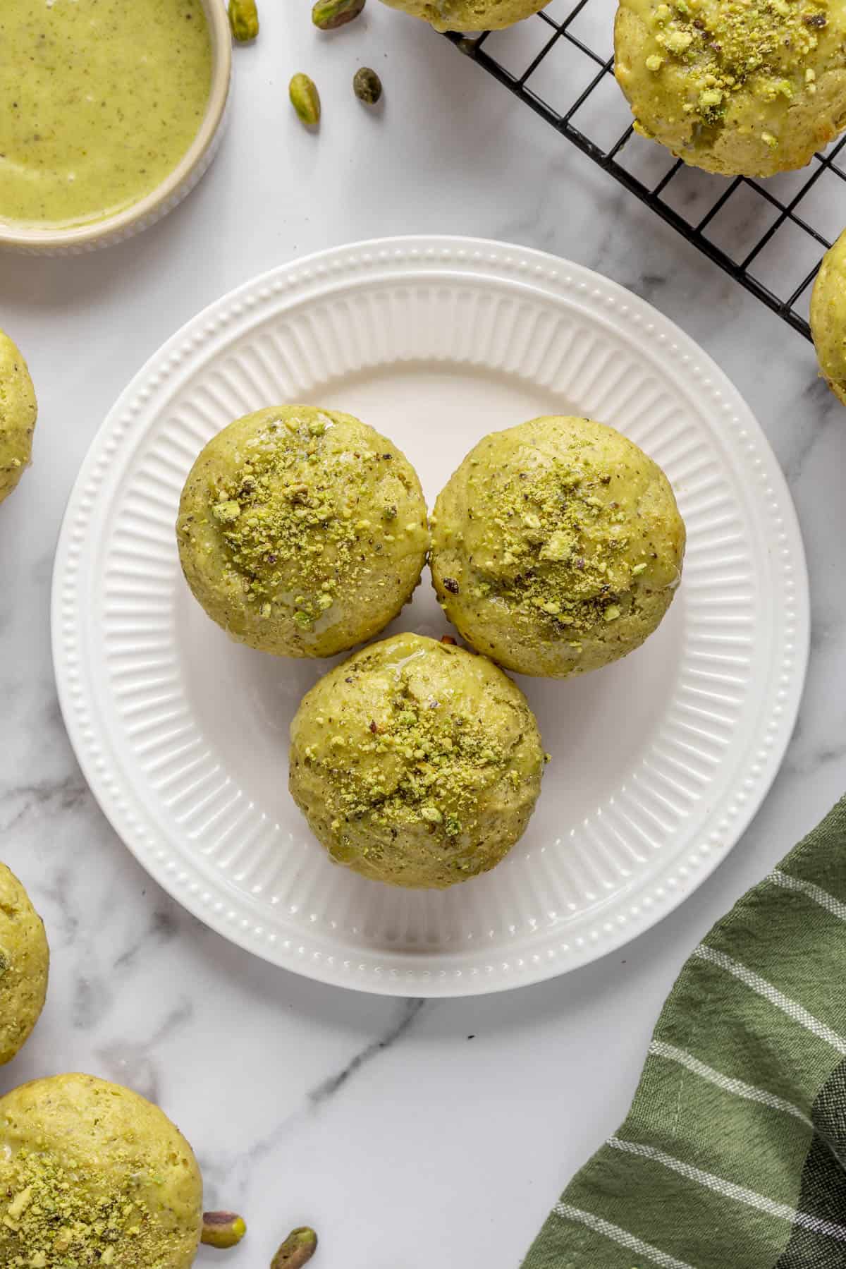 Pistachio muffins topped with glaze and crushed pistachios on a plate with more muffins and glaze nearby.