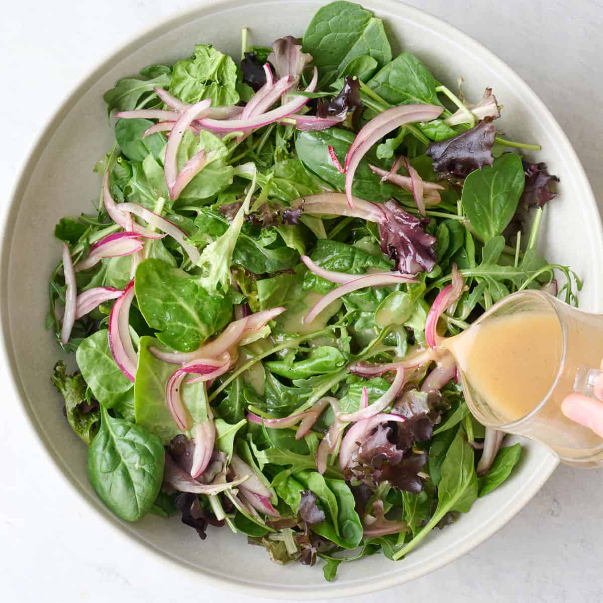 Pouring apple cider vinaigrette dressing over mixed greens in a bowl.
