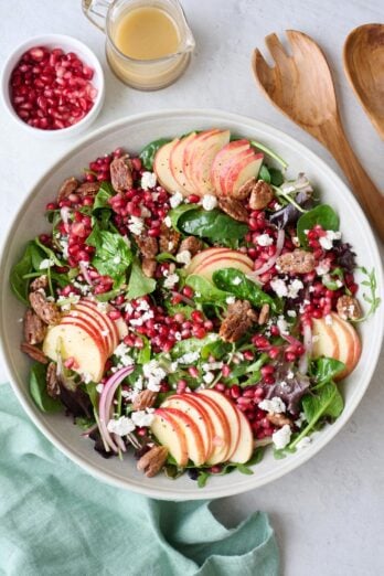 Pomegranate salad recipe in a large salad bowl topped with sliced red onions, pomegranate arils, candied pecans, crumbled goat cheese, and thinly sliced apples.