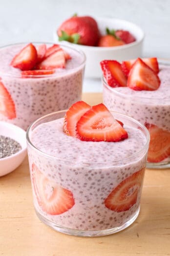 Strawberry chia pudding in 3 mini glasses with a few sliced strawberries against the glass and also on top, dish of fresh berries and dish chia seeds nearby.