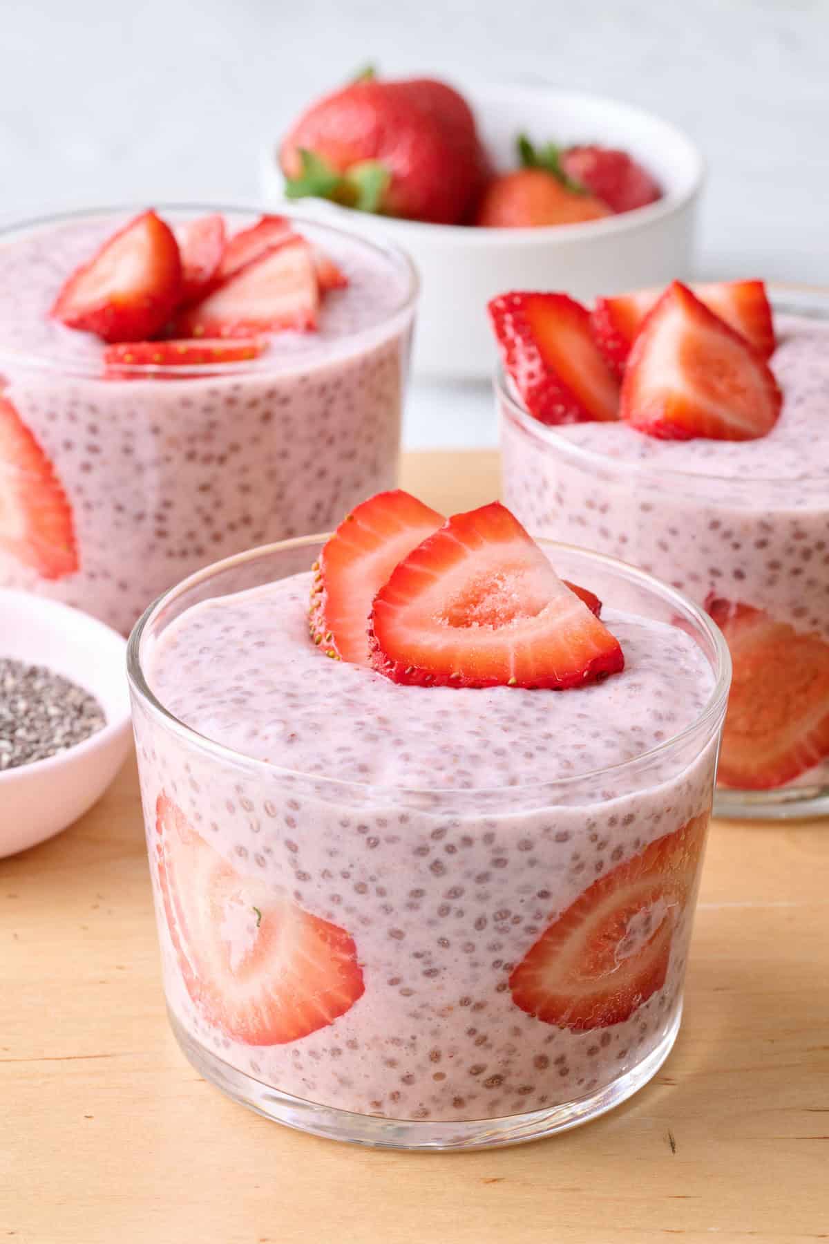 Strawberry chia pudding in 3 mini glasses with a few sliced strawberries against the glass and also on top, dish of fresh berries and dish chia seeds nearby.