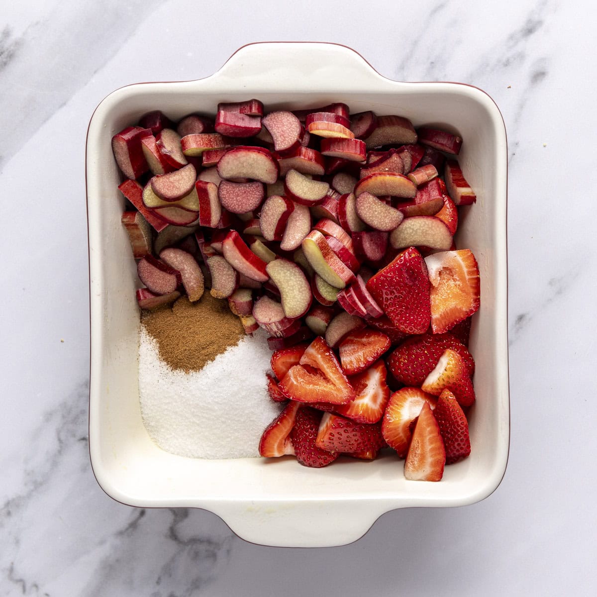 Chopped rhubarb, strawberries, sugar, and cinnamon in a square baking dish before tossing together.
