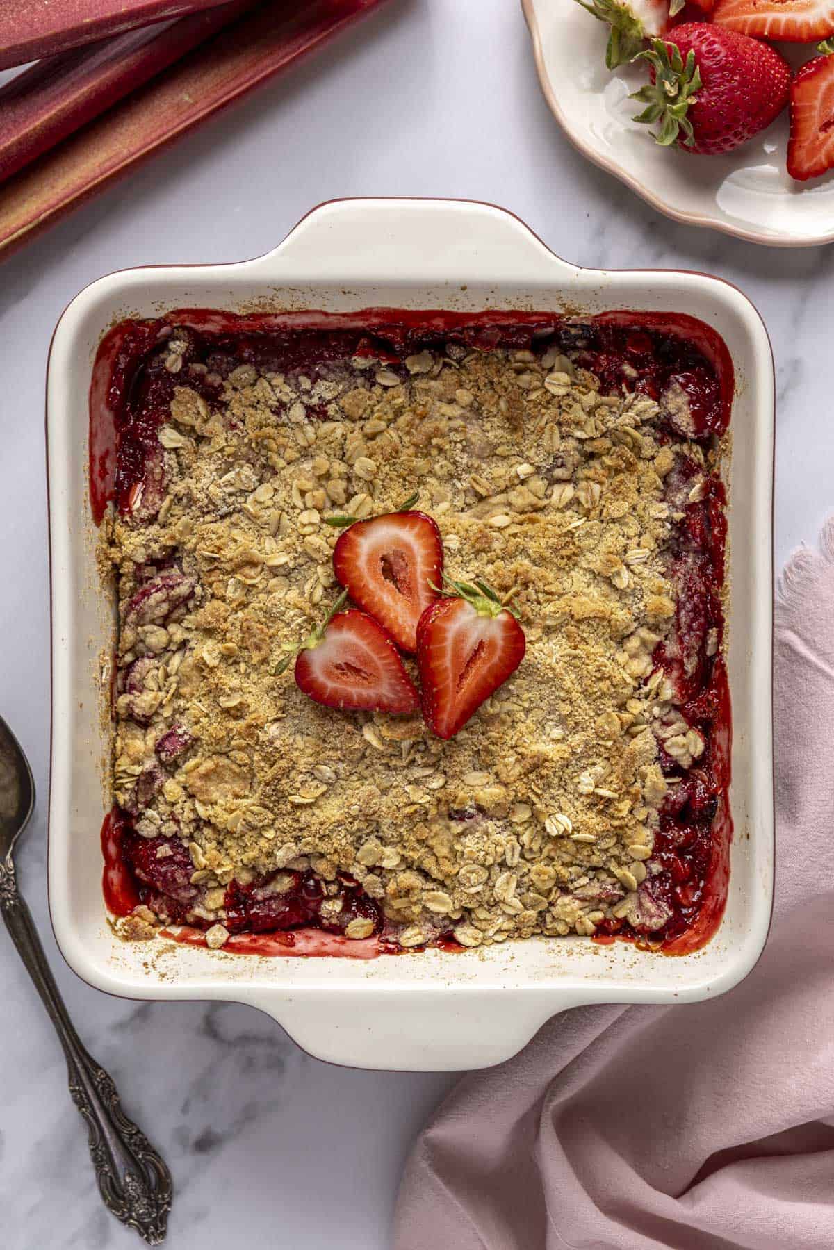 Strawberry rhubarb crisp in baking dish a few slices of fresh strawberries on top and with extra fruit and serving spoon nearby.