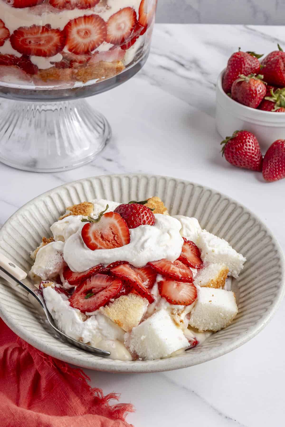 Serving of homemade strawberry trifle in a bowl with trifle dish and fresh strawberries nearby.