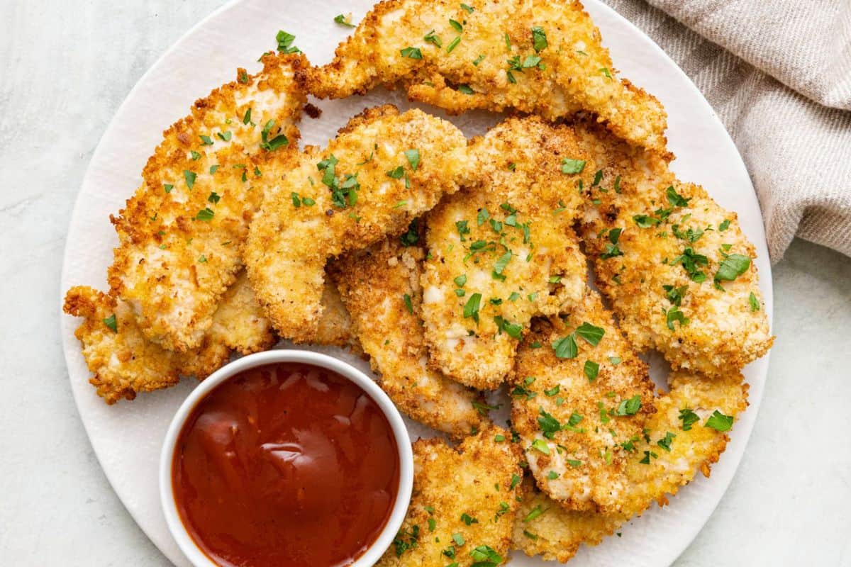 Crispy air fryer chicken tenders on a plate with a small dish of ketchup for dipping.