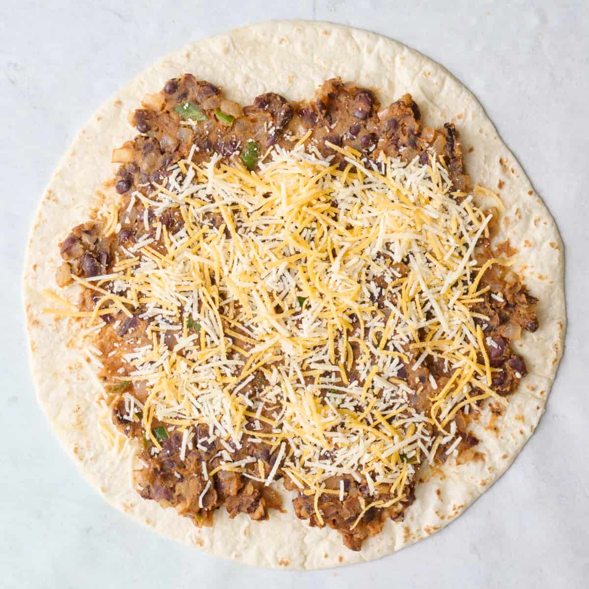 Tortilla on a flat surface with bean mixture on half with cheese sprinkled on top.