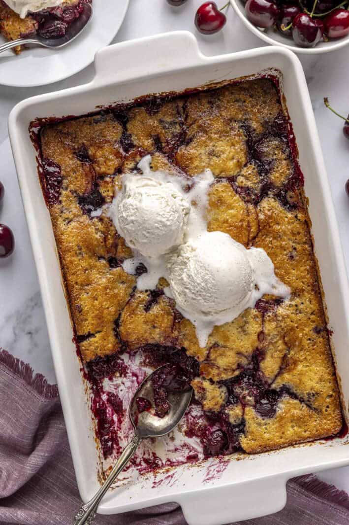 Homemade cherry cobbler in a baking dish with a serving removed and two scoops of vanilla ice cream on top.