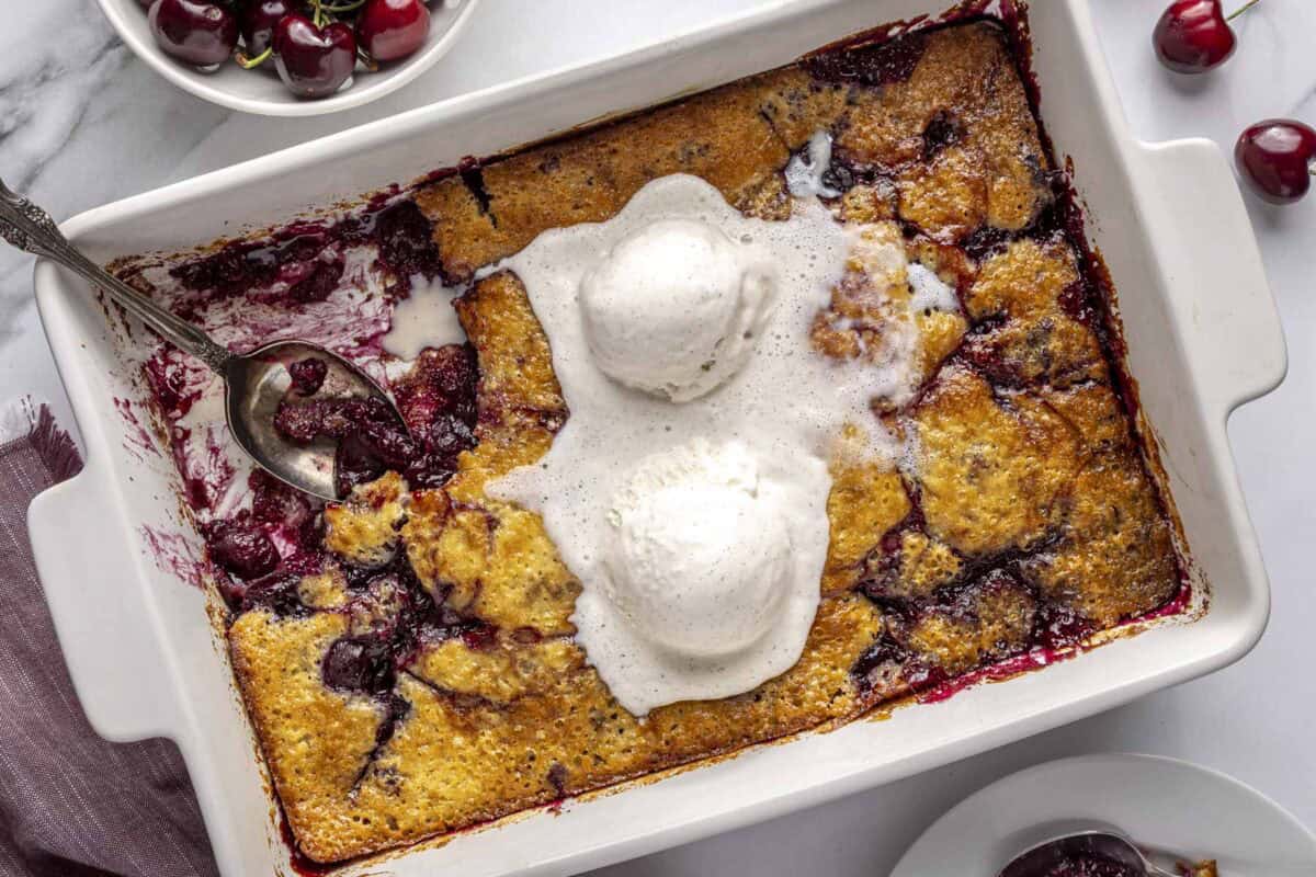 Homemade cherry cobbler in a baking dish with a serving removed and two scoops of vanilla ice cream on top.