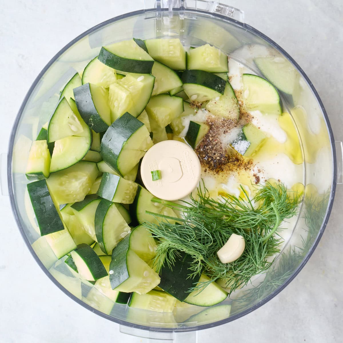 Chopped cucumbers, dill, garlic, yogurt, oil, and seasonings in a food processor before processing.