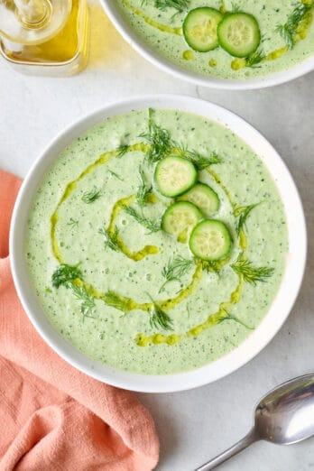 Two bowls of cucumber soup with focus on one, olive oil drizzled in a circular pattern on top and garnished with thinly sliced cucumbers and fresh dill, spoon resting nearby.
