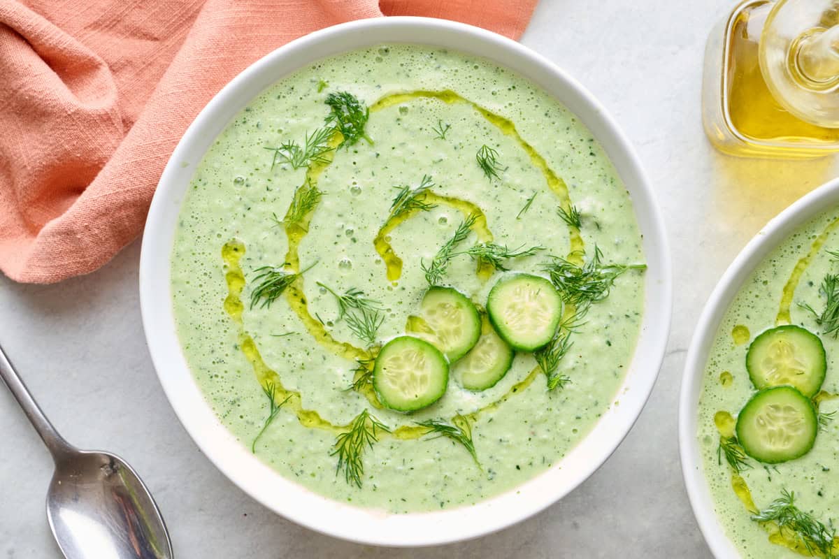 Two bowls of cucumber soup with focus on one, olive oil drizzled in a circular pattern on top and garnished with thinly sliced cucumbers and fresh dill, spoon resting nearby.