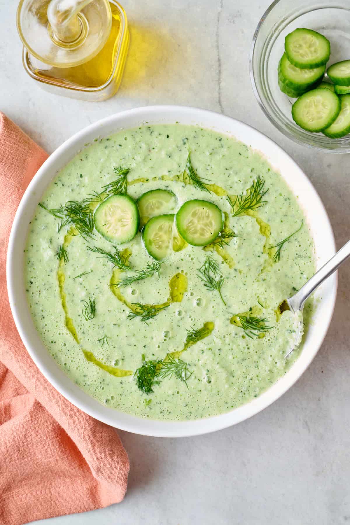 Creamy cucumber soup in a bowl with a spoon dipped inside, garnished with extra slices of cucumbers and a drizzle of olive oil.