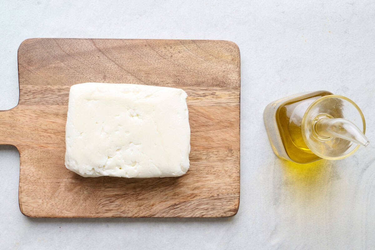Ingredient for recipe: block of halloumi on a small cutting board and bottle of olive oil.