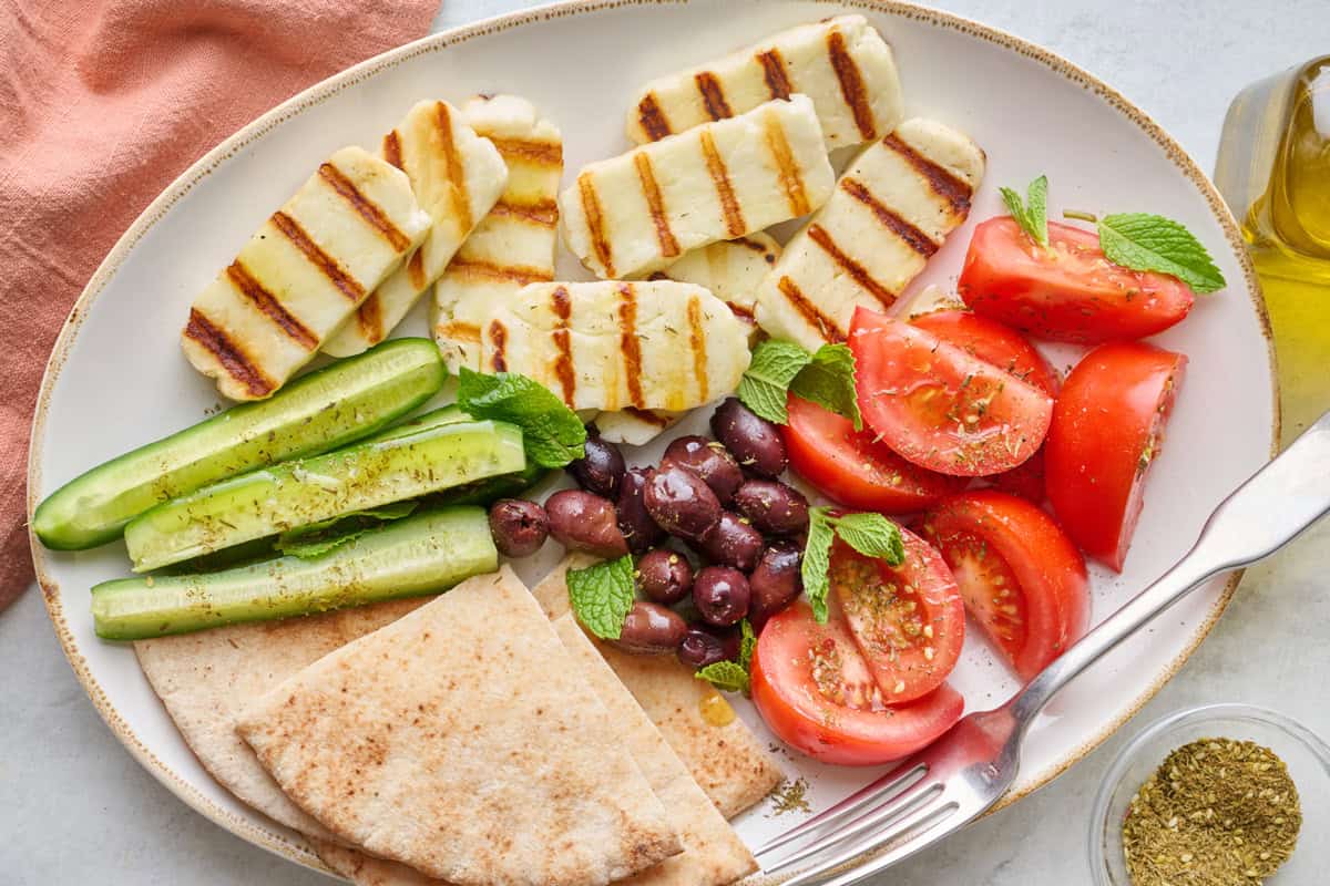 Grilled halloumi on a large oval platter with sliced tomatoes, sliced cucumbers, fresh mint, zaatar, and a drizzle of olive oil, greek pita and serving fork nearby.