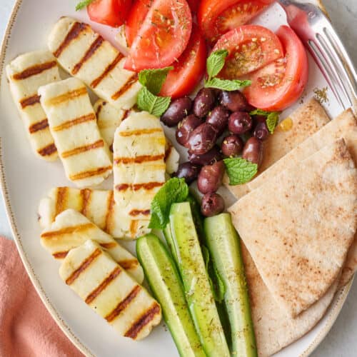 Grilled halloumi recipe on platter with fresh veggies, olives, and pita.
