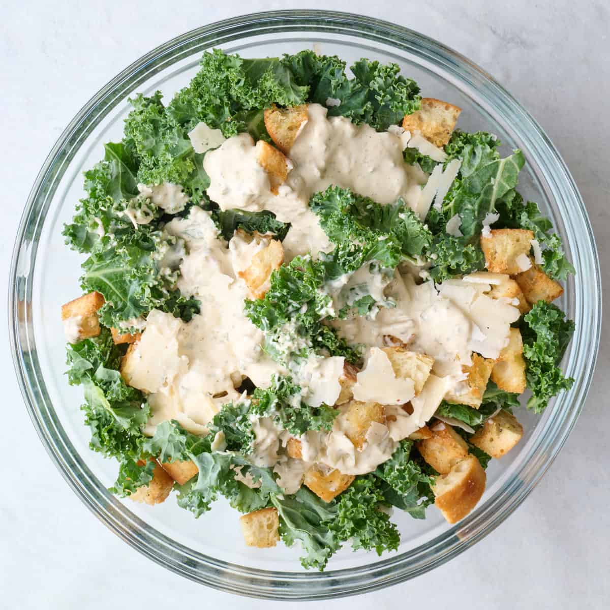 Kale, shaved parmesan and croutons in a bowl with dressing drizzled on top.