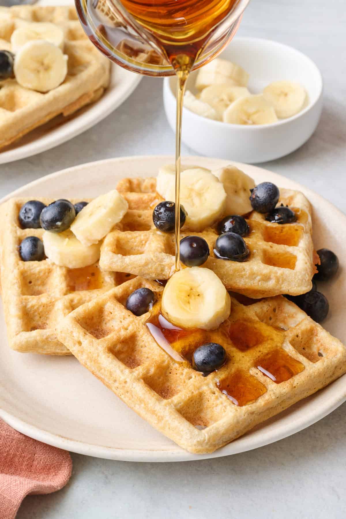Syrup being poured over a plate of homemade oatmeal waffles topped with fruit.