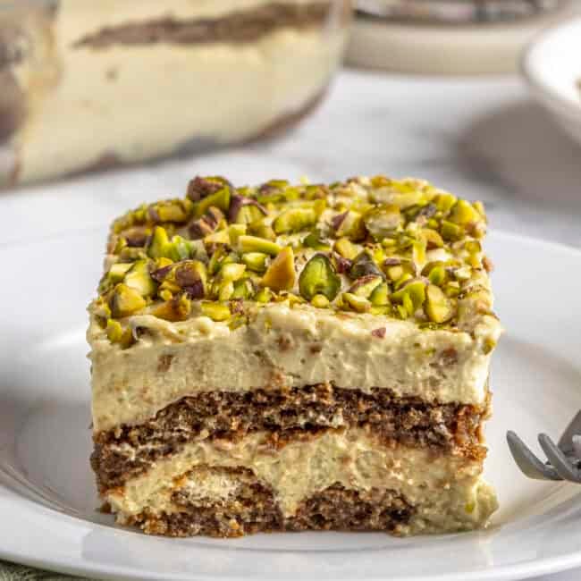No bake pistachio tiramisu slice on a plate showing layers of pistachio mascarpone and espresso dipped lady fingers, topped with crushed pistachios.