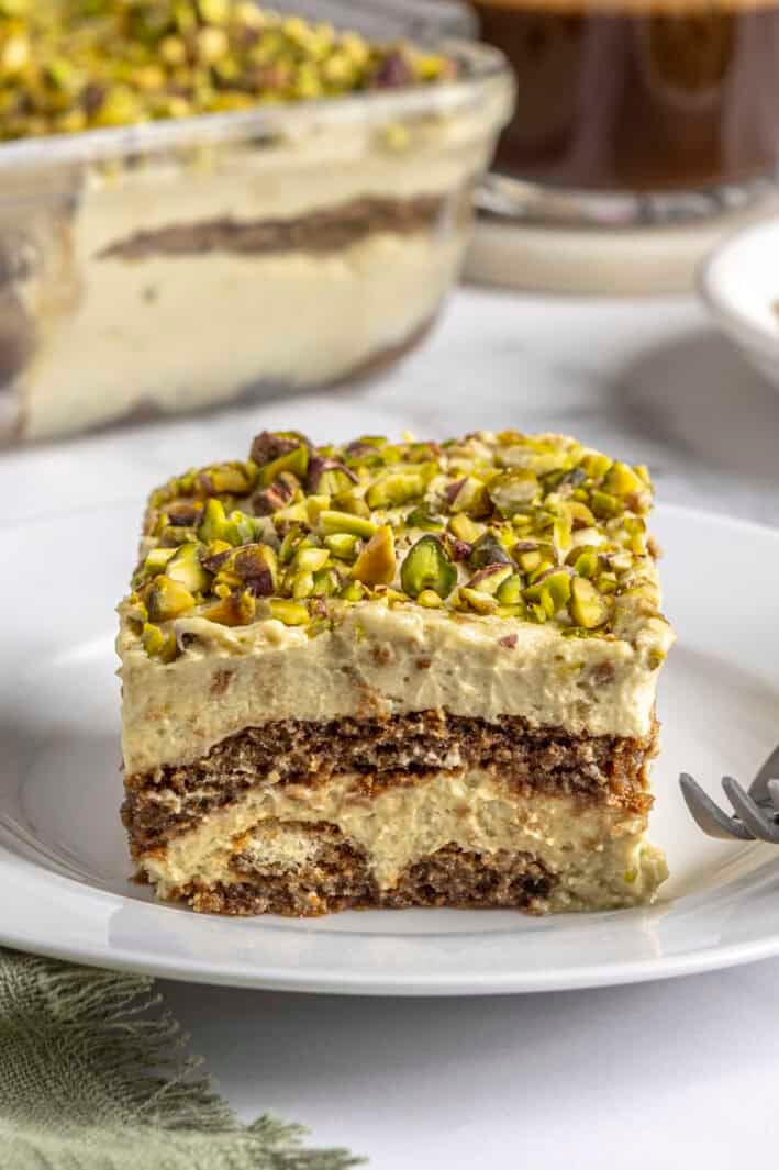 No bake pistachio tiramisu slice on a plate showing layers of pistachio mascarpone and espresso dipped lady fingers, topped with crushed pistachios.