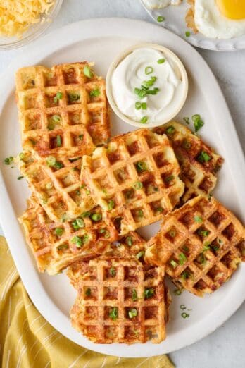 Crispy potato waffles on a platter garnished with chopped green onions, with a small dish of sour cream and small plates of cooked eggs nearby.