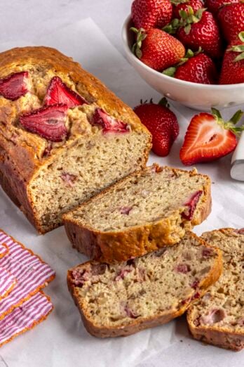 Strawberry banana bread loaf on parchment paper with a few slices cut away and bowl of fresh strawberries nearby.
