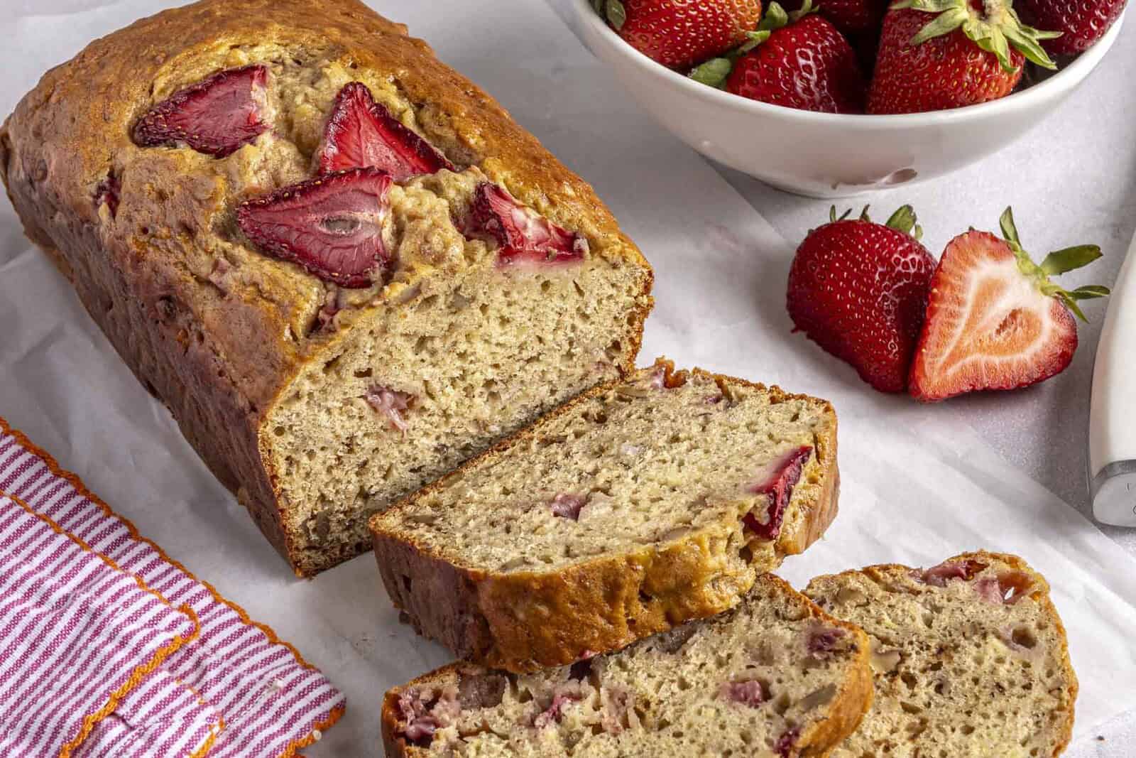 Strawberry banana bread loaf on parchment paper with a few slices cut away and bowl of fresh strawberries nearby.