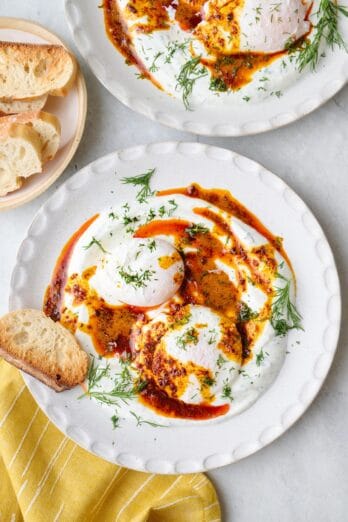Two bowls of Turkish eggs served over yogurt, topped with hot chili butter and garnished with fresh dill, crust bread nearby.