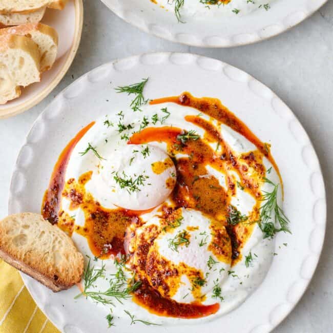 Two bowls of Turkish eggs served over yogurt, topped with hot chili butter and garnished with fresh dill, crust bread nearby.