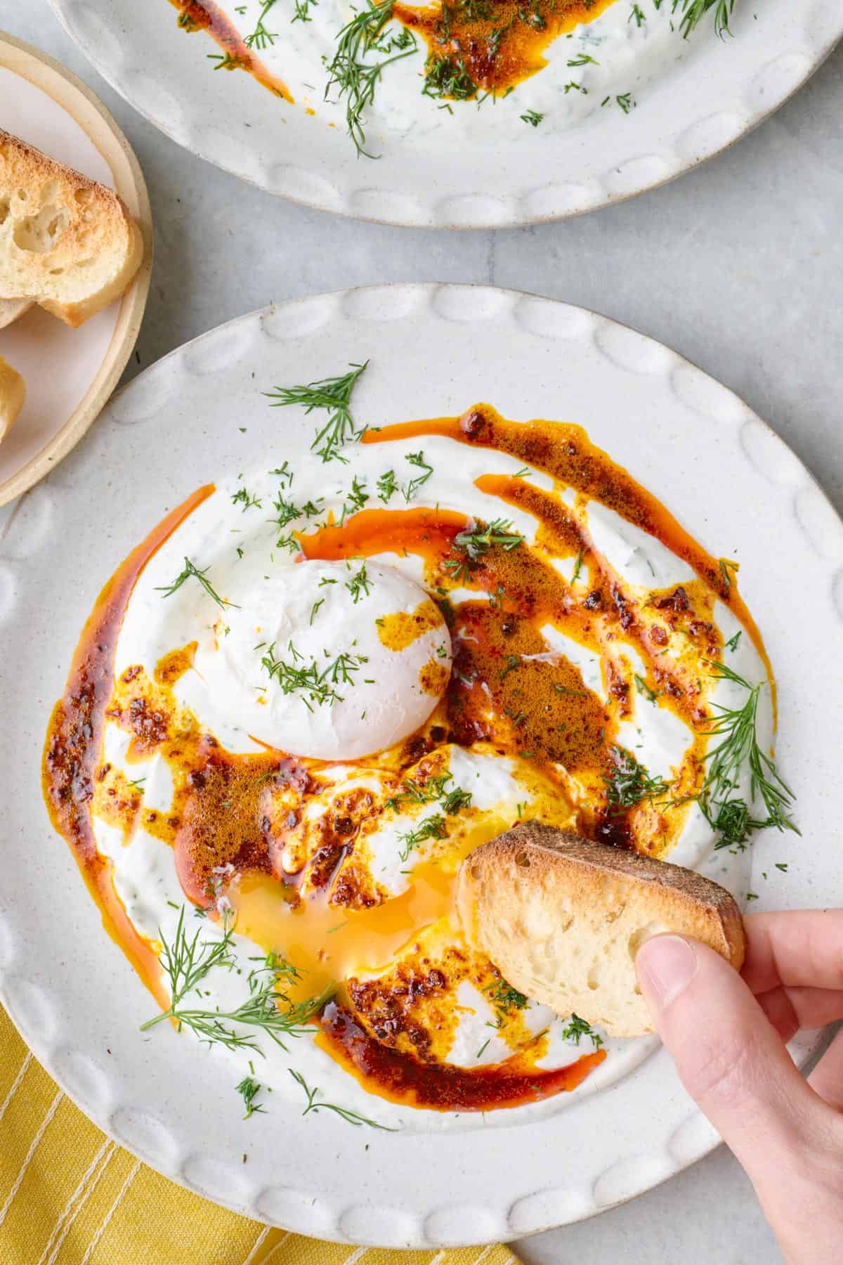 Two bowls of Turkish eggs served over yogurt, topped with hot chili butter with crusty bread being dipped into runny yolk.