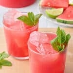 Two glasses filled with ice and watermelon juice with pitcher nearby and mint leaves on top, extra watermelon (triangles) nearby.