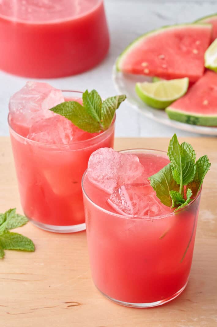 Two glasses filled with ice and watermelon juice with pitcher nearby and mint leaves on top, extra watermelon (triangles) nearby.