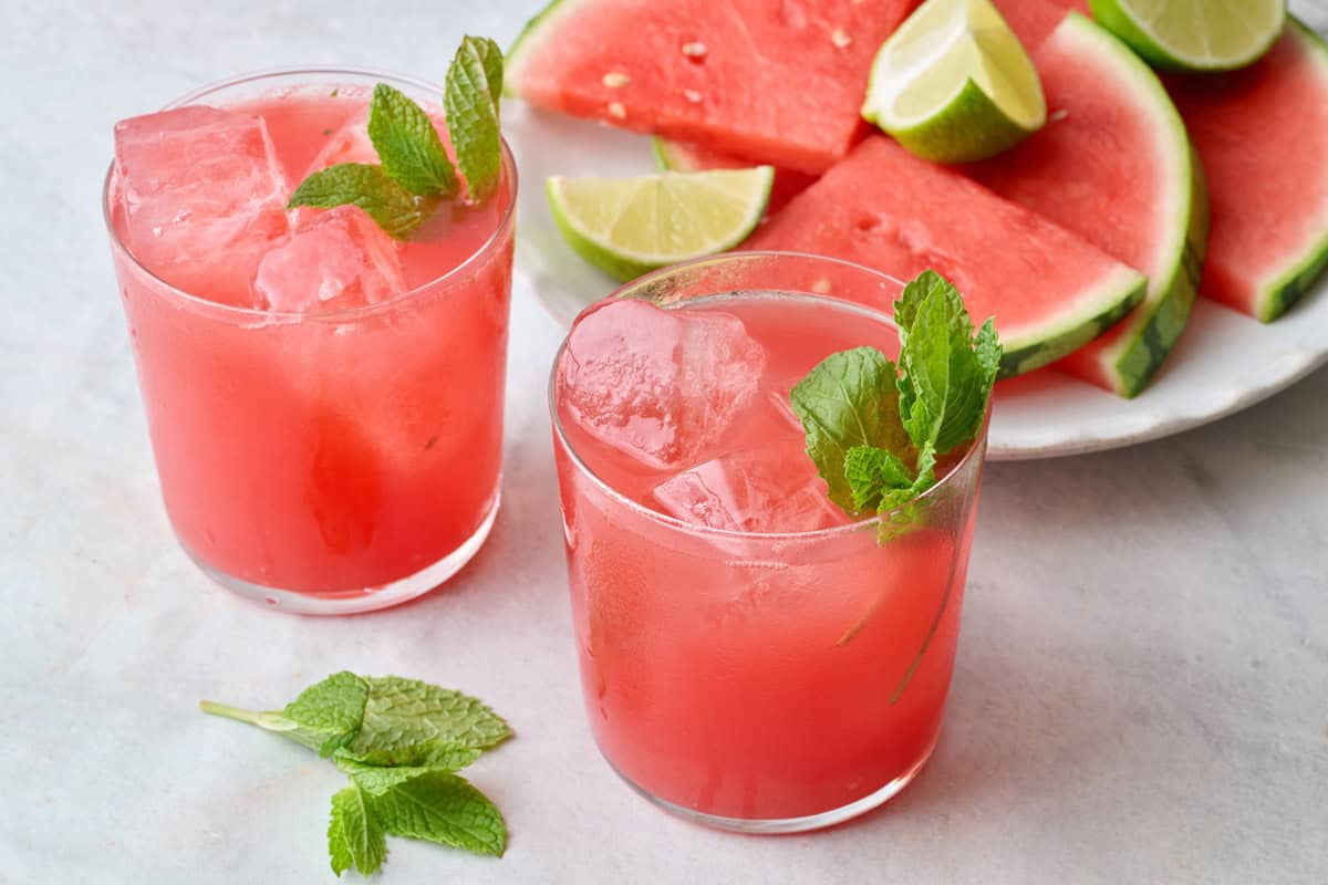 Two glasses filled with ice and watermelon juice with pitcher nearby and mint leaves on top, extra watermelon (triangles) nearby.