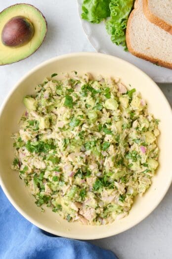 Avocado tuna salad in a serving bowl with a few slices of bread, leafy lettuce and half avocado nearby.