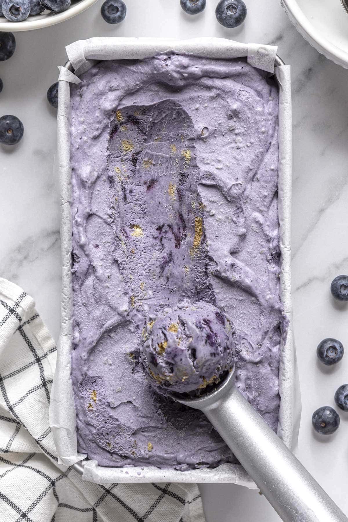 Blueberry ice cream with graham crackers in a dish with ice cream scoop resting on top with some ice cream on it.