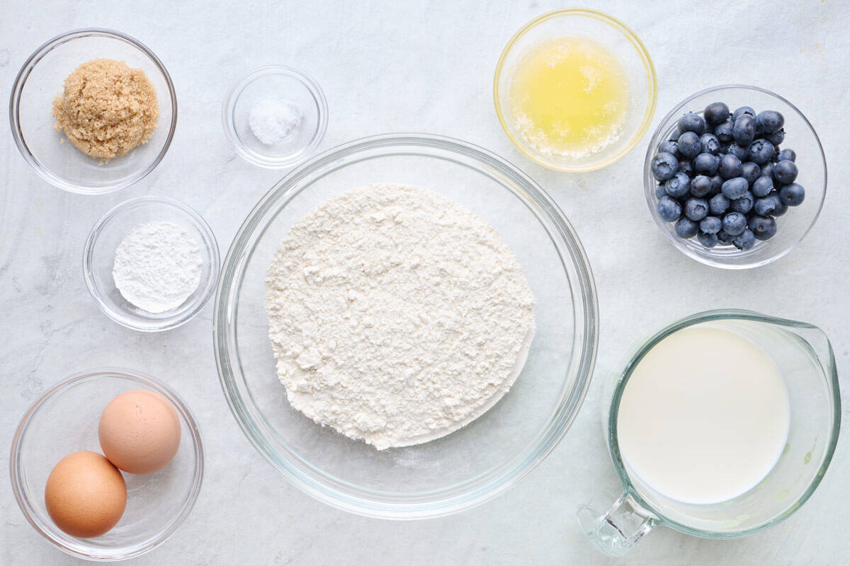 Ingredients for recipe in individual bowls: brown sugar, baking powder, salt, eggs, flour, melted butter, blueberries and milk.