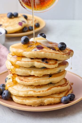 Stack of blueberry pancakes topped with a pat of slightly melted butter and fresh blueberries, with maple syrup being poured on top.