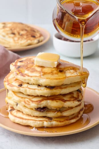 Stack of chocolate chip pancakes on a plate with butter on top and syrup being poured over them.