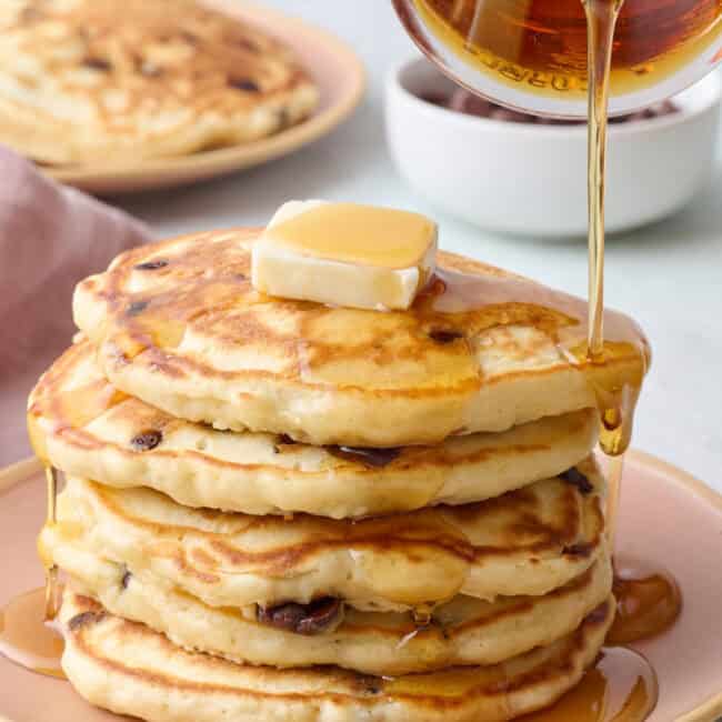 Stack of chocolate chip pancakes on a plate with butter on top and syrup being poured over them.