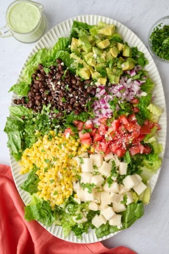 Chopped Mexican salad on a large platter with lettuce, black beans, corn, jicama, tomatoes, red onions, and diced avocado with dressing nearby.