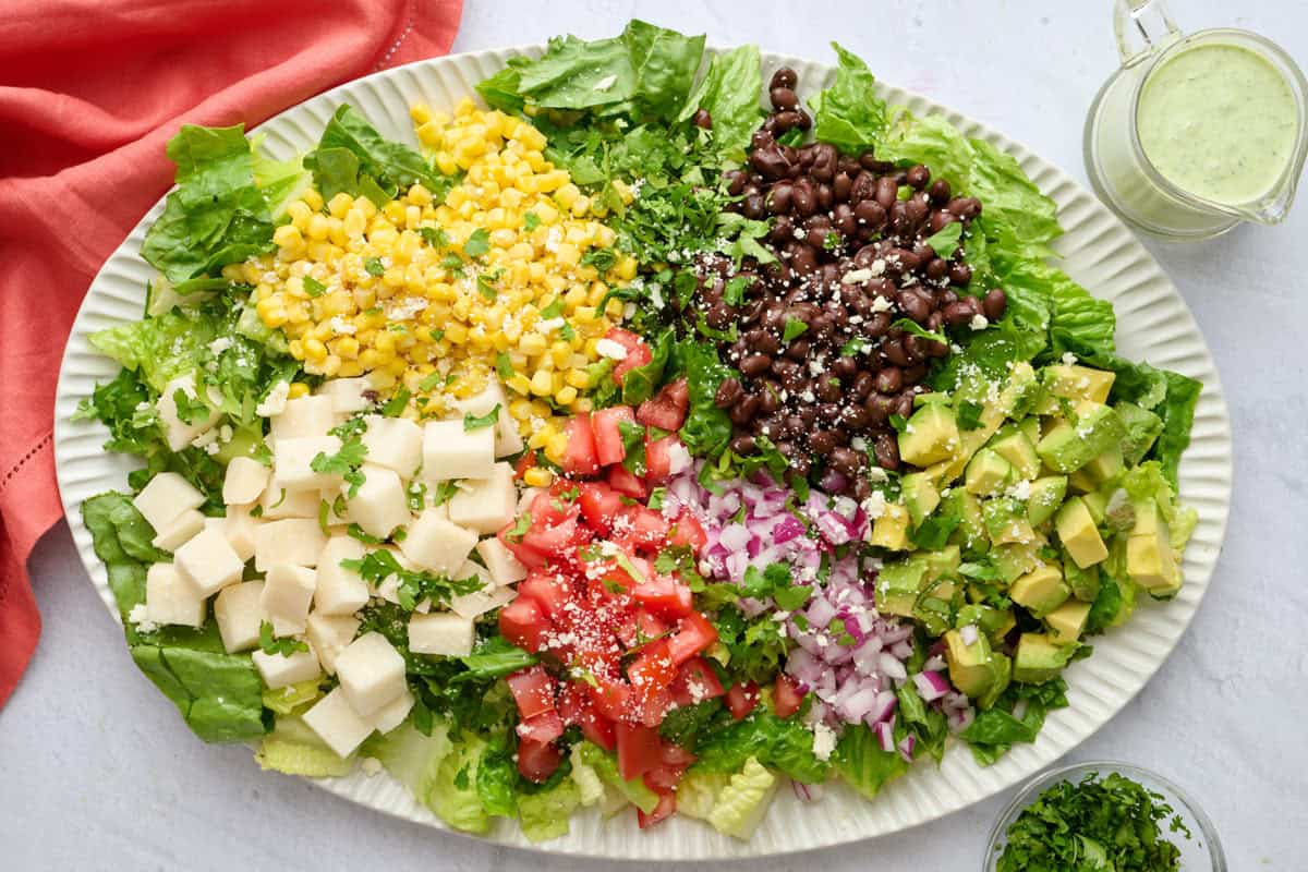 Chopped Mexican salad on a large platter with lettuce, black beans, corn, jicama, tomatoes, red onions, and diced avocado with dressing nearby.