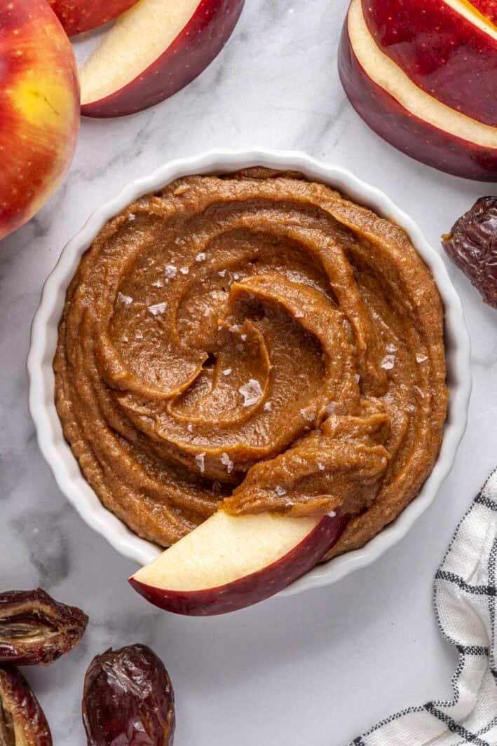 Date caramel in a small dish, top swirled, with extra dates and whole and sliced apples nearby.
