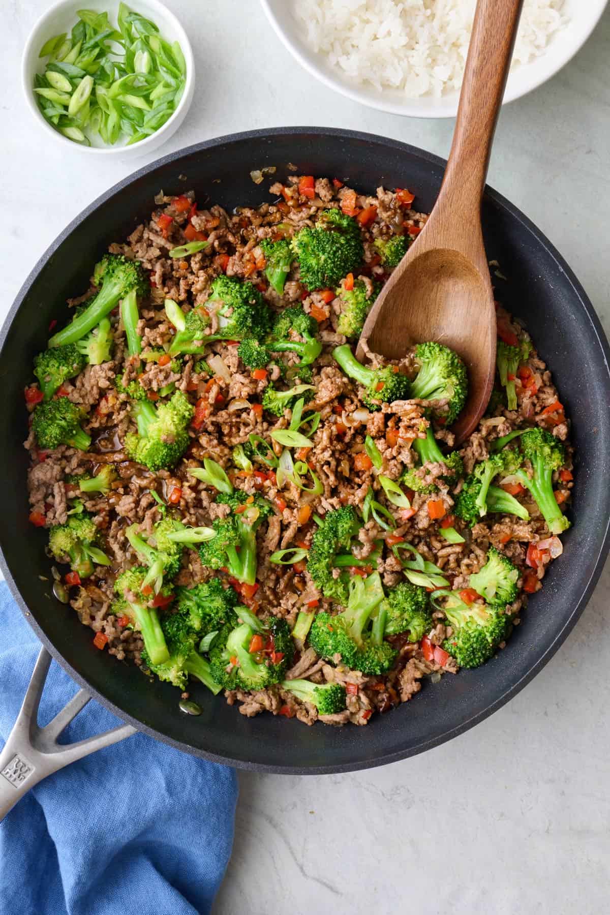Ground beef stir fry in a skillet with wooden serving spoon, topped with green onions with serving bowl of white rice nearby.