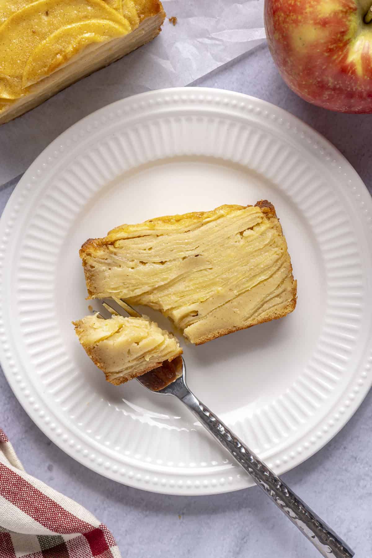 Slice of invisible apple cake on a plate with a bite on a fork resting on plate.
