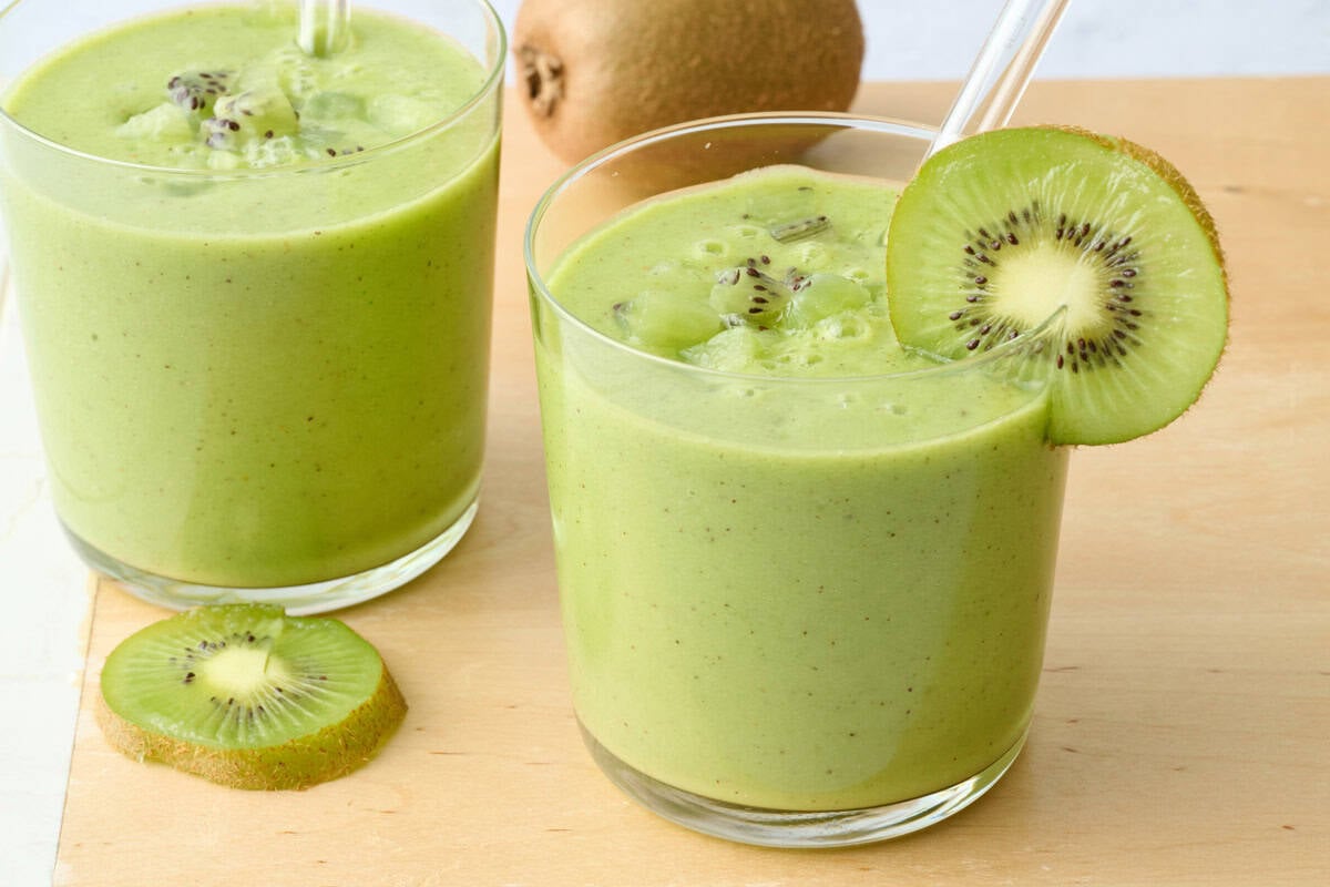 2 glasses of kiwi smoothie with a few kiwi chunks on top and slice kiwi on rim, clear reusable straws dipped inside.