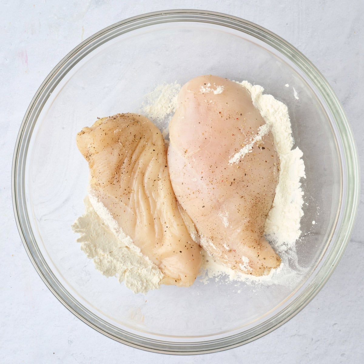 Seasoned chicken breasts in top of a bowl of flour.