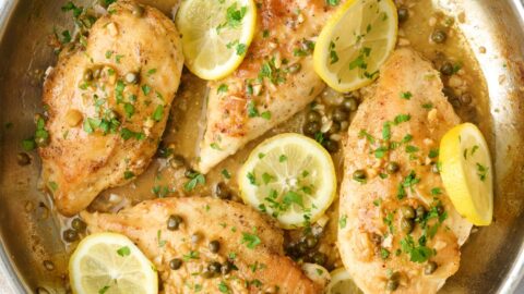 Lemon chicken piccata in skillet with capers and sauce spooned over chicken, garnished with chopped parsley and fresh sliced lemons on top.