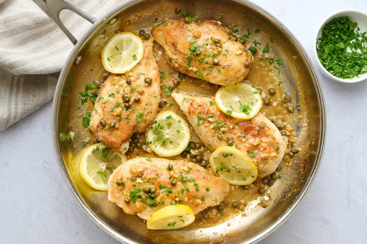 Lemon chicken piccata in skillet with capers and sauce spooned over chicken, garnished with chopped parsley and fresh sliced lemons on top.