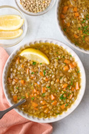 Two bowls of soup with focus on one, garnished with parsley and lemon wedge, spoon dipped inside, extra garnishes or small dish of dried lentils nearby.