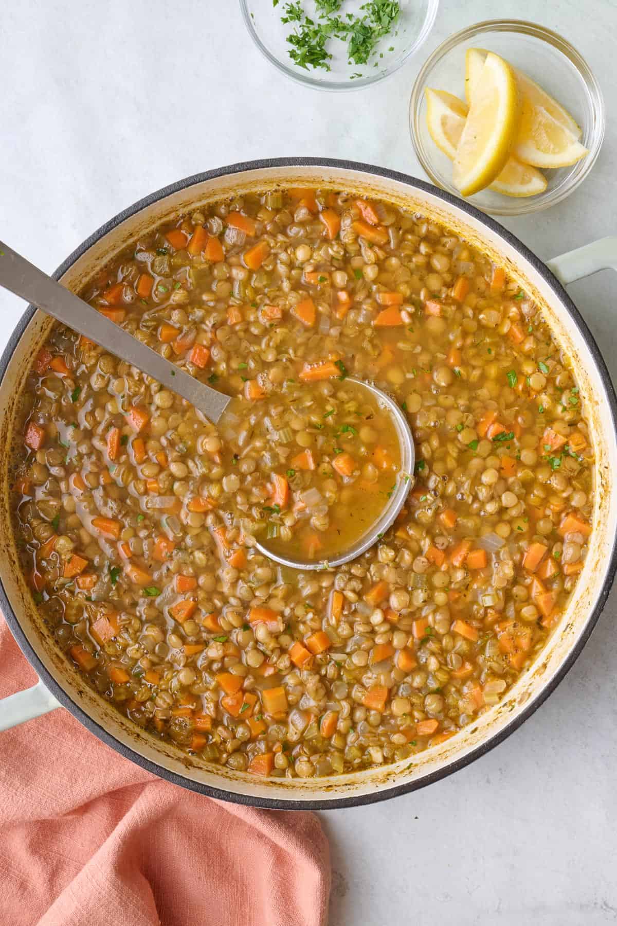 Pot of lentil soup with ladle dipped inside, garnished with fresh parsley with extra lemon wedges nearby.